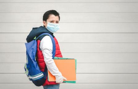Child carrying a backpack and books while wearing a mask and gloves.
